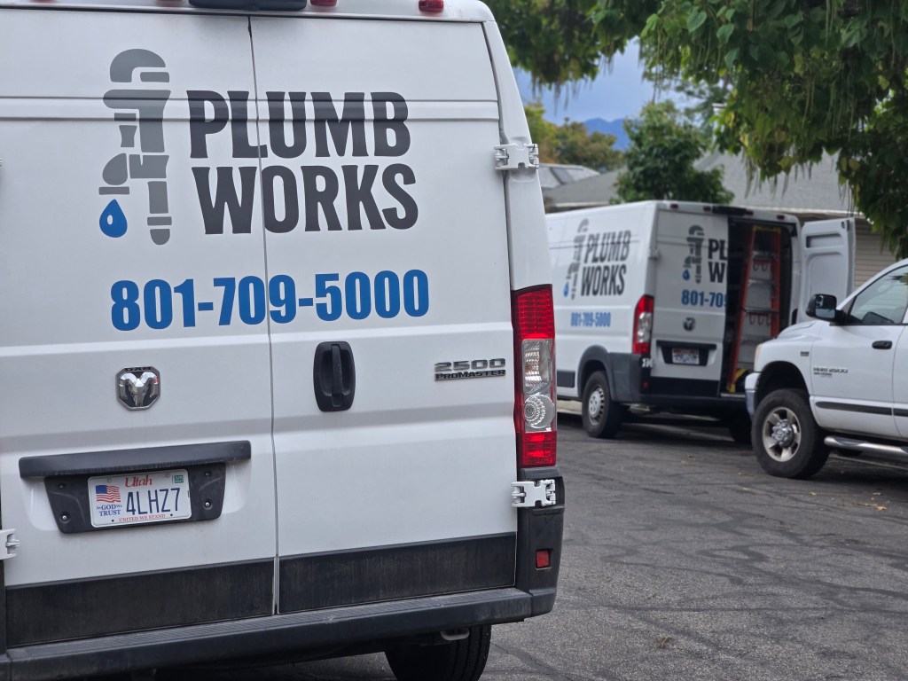 Two service vans parked on a residential street, one prominently displaying the logo and contact information for 'Plumb Works.'